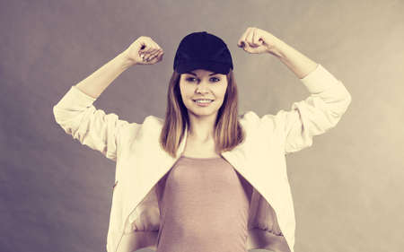 Young sporty woman wearing cap and sportswear showing her arm muscles enjoying workout results. Studio shot on grey backgroundの写真素材