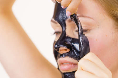 Young woman removing carbo black peel-off mask from her face, on white. Teen girl taking care of oily skin, cleaning the pores. Beauty treatment. Skincare.の写真素材