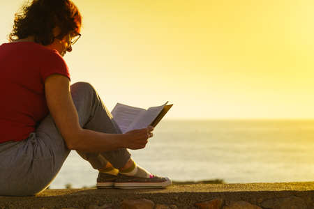 Mature tourist woman relaxing on coast with book, enjoy fresh air in sunset time. Reading on holidays. Relax time.の写真素材