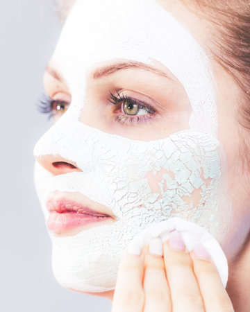 Young woman with white dried mud mask on face being removing cosmetic with cotton swab. Teen girl taking care of oily skin. Beauty treatment. Skincare.の写真素材