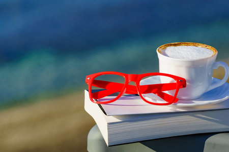 Spiritual healing, psyche silence. Coffee cup, book and red glasses against coast. Reading on vacation. Relaxation on holidays. Mental health break.の写真素材