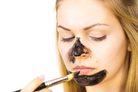 Young woman being apply with brush purifying charcoal mask, black cosmetic to her face. Female taking care of oily skin. Beauty treatment. Skincare.の写真素材