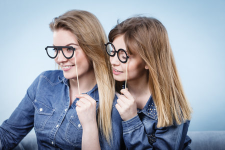 Two happy women holding fake eyeglasses on stick having fun fooling around wearing jeans shirts. Photo and carnival funny accessories concept.の写真素材