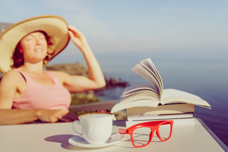 Reading on holidays. Mature tourist woman relaxing on coast, read book, enjoy sea view from cliff. Focus on coffee cup and glasses.の写真素材