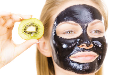 Young woman with carbo black peel-off mask on her face holding kiwi fruit halves, on white. Beauty treatment. Skincare.の写真素材