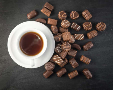 Beautiful Creative Chocolate Sweets on Natural Black Stone with White Coffee Cup. Background. Scattered Chocolates Top View and Flat Lay with Place for Textの写真素材