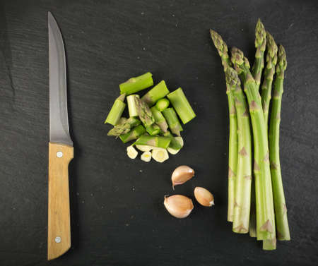 Chopped Raw Garden Asparagus with Garlic and Knife Ready for Frying Top View. Fresh Green Spring Vegetables on Stone Background.の写真素材