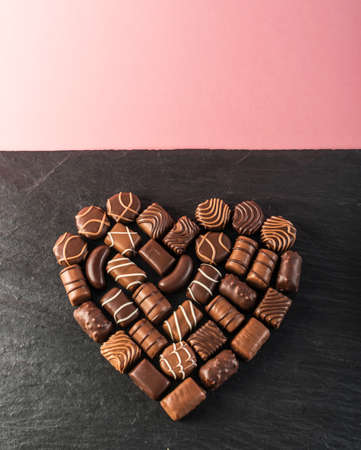 Beautiful Creative Chocolate Heart Shape Made with Various Types of Pralines Over an Black Stone Background. Sweet Chocolates with Pink Backdropの写真素材