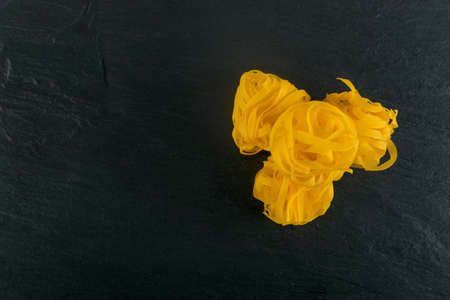 Raw yellow italian pasta fettuccine, fettuccelle or tagliatelle close up. Egg homemade dry ribbon noodles, long rolled macaroni or uncooked spaghetti on black stone background top viewの写真素材