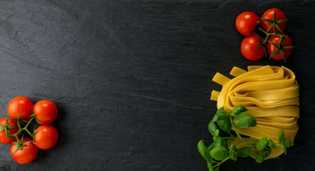 Raw yellow italian pasta pappardelle, fettuccine or tagliatelle close up. Egg homemade dry ribbon noodles, long rolled macaroni or uncooked spaghetti with tomatoes and basil top viewの写真素材