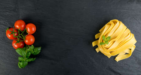 Raw yellow italian pasta pappardelle, fettuccine or tagliatelle close up. Egg homemade dry ribbon noodles, long rolled macaroni or uncooked spaghetti with tomatoes and basil top viewの写真素材