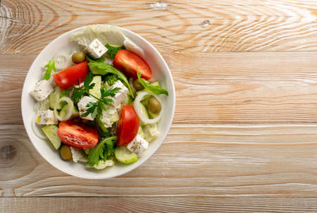Greek salad or horiatiki with large pieces of tomatoes, cucumbers, feta cheese and olives in white bowl top view, copyspace. Village salad with diced mozzarella, arugula, parsley, spices and oilの写真素材