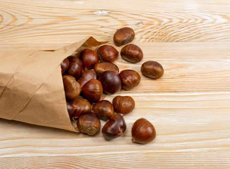 Edible sweet whole chestnuts in paper bag on wood table background top view. Raw healthy delicious chestnut, autumn and christmas european veggie foodの写真素材
