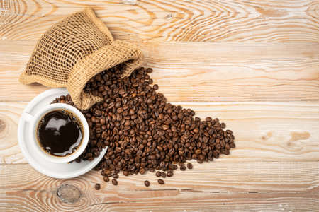Hot espresso cup and coffee beans in burlap bag on wooden background. White coffeecup or mug, whole roasted arabica coffea bean in natural jute rustic sack with copyspaceの写真素材