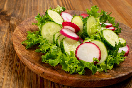 Spring homemade radish salad with fresh cucumber and greens on wooden plate. Simple green rustic salat with sliced radishes, cucumbers, escarole and lettuceの写真素材