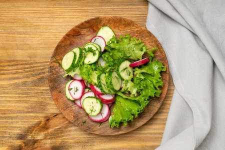 Spring homemade radish salad with fresh cucumber and greens on wooden plate top view. Simple green rustic salat with sliced radishes, cucumbers, escarole and lettuceの写真素材
