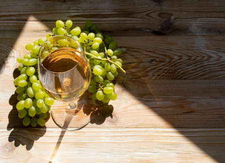 White wine glass on ripe bunch of grapes background in sunny day. Wineglass with golden grape sparkling drink on wooden rustic winery table top viewの写真素材