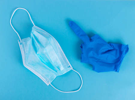 Medical mask and used medical gloves as if showing the middle finger on blue background. Surgical masks, disposable medic clothing item, hospital equipment, corona virus face protection conceptの写真素材