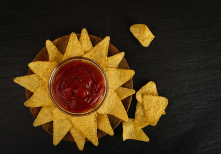 Triangle corn chips with red sauce on dark rustic background. Mexican nachos chips for nacho tortilla, maize snack, corn crisps or totopos with salsa dip in vintage country style top viewの写真素材