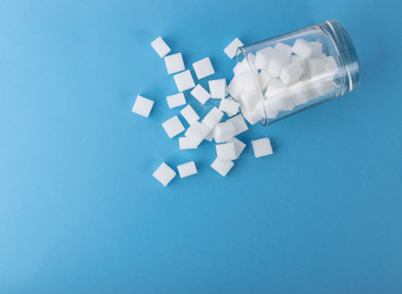 Spilled white sugar cubes on blue background. Unrefined cane sugar cube in drinking glass top view