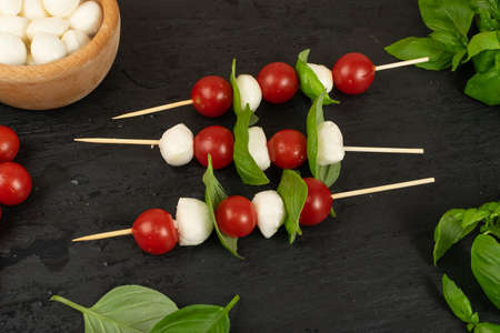 Mozzarella and cherry tomatoes on skewers on black slate background. Italian caprese salad with mozarella cheese balls, tomato and basil top viewの写真素材
