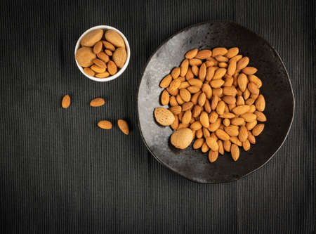 Heap of almond in black bowl on dark background with copy space. Scattered almonds with shell and peeled nuts top viewの写真素材