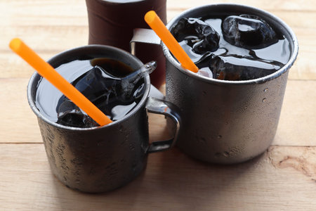 Two cups of black coffee with ice cubes and straws on wooden table