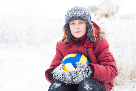 Portrait of a boy in a red jacket sitting in the snow with a volleyball ball