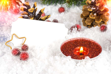 Christmas decoration with a red candle in the snow and pine cones in the background and red berries, one golden star and a label for labeling in the foregroundの写真素材
