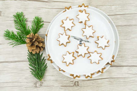 A white plate with Christmas biscuits, cinnamon stars with frosting on wooden background, on the left is a pine branch and a pine coneの写真素材