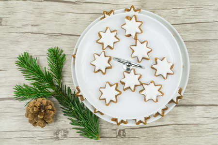 A white plate with Christmas biscuits, cinnamon stars with frosting on wooden background, on the left is a pine branch and a pine coneの写真素材