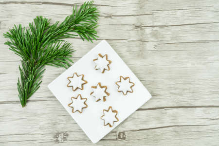 A white square plate with Christmas biscuits, cinnamon stars with frosting on wooden background, on the left is a pine branch, Christmas time, copyspace,の写真素材