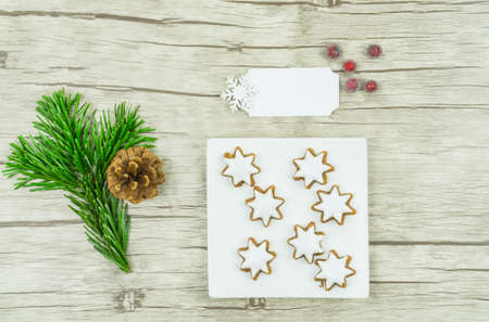 A white square plate with Christmas biscuits, cinnamon stars with frosting on wooden background, on the left is a pine branch with a pine cone, berries and to sign inscribable, Christmas timeの写真素材