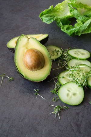 A halved avocado with core, many slices of a cucumber salad, a lettuce leaf and cress, background black slateの写真素材