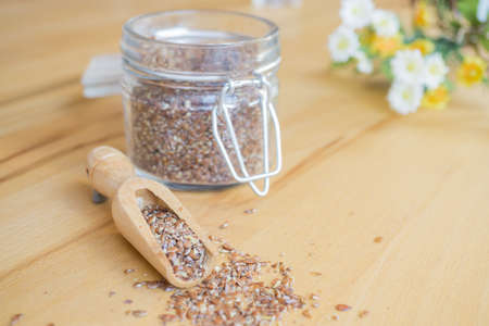 A glass jar with crushed linseed, beside it a small wooden spoon with linseed and some summer flowers on the upper right edge of the picture, background of woodの写真素材