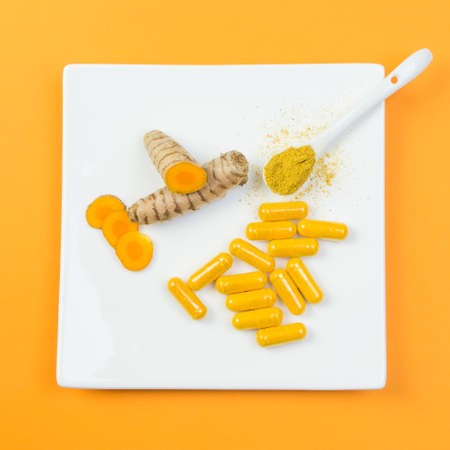 Turmeric root with yellow capsules and a spoonful of curcuma powder on a white plate with orange backgroundの写真素材
