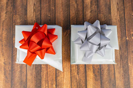 Two white-wrapped gifts on a wooden table, one with a red bow and the other with a silver bowの写真素材