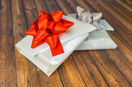 Two white-wrapped gifts on a wooden table, one with a red bow and the other with a silver bow