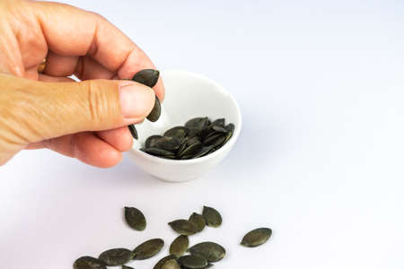 A woman holds pumpkin seeds in her hand, in the background is a white porcelain bowl with pumpkin seeds, copy spaceの写真素材