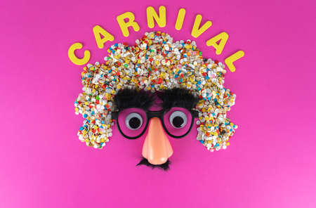 Funny face mask with big eyes and confetti hair, pink background with copy space and the word carnival in yellow lettersの写真素材