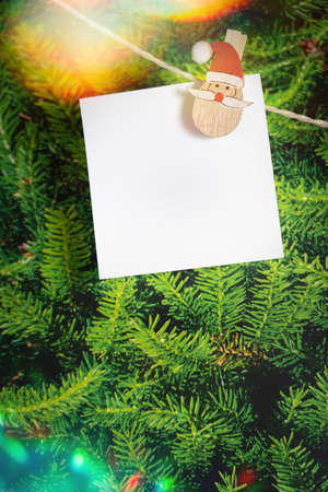 White blank sticky note with a Santa Claus head attached to a cord, in the background a fir tree, copy spaceの写真素材