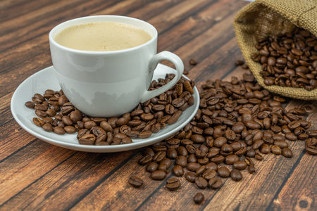A cup of creamy coffee with roasted coffee beans next to it, wooden backgroundの写真素材