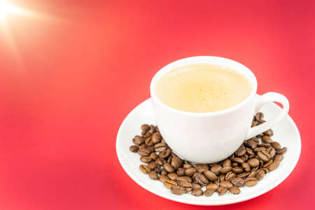 A cup of creamy coffee with roasted coffee beans on the saucer, red background and copy spaceの写真素材