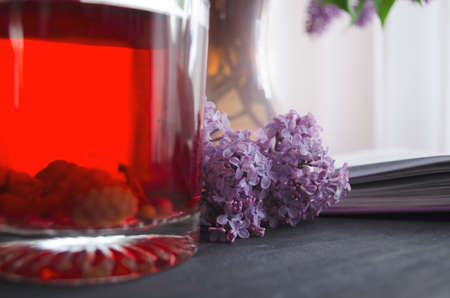 Still life with a bouquet of lilacs and compote of berries. A glass of compote, a jug, a bouquet, a book and glasses on a dark wooden table. Still life near the window.の写真素材