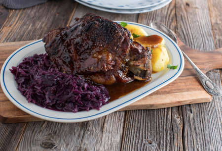 Homemade holiday roast with roasted turkey shank, red cabbage, boiled potatoes and delicious gravy. Served on a plate isolated on wooden table background with copy spaceの写真素材