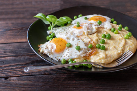 Fried eggs sunny side up with creamy polenta and green peas on dark wooden background. Closeup and front viewの写真素材