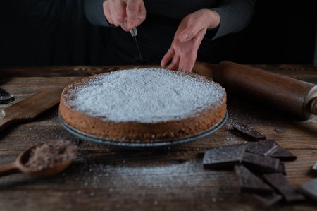Woman with older hands is ready to slice a freshly baked chocolate cake on rustic and dark wooden table background with old fashioned kitchen utensils.の写真素材