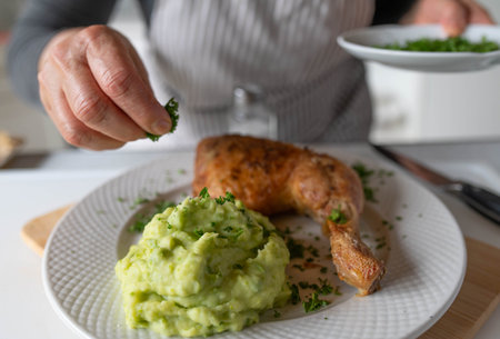 Woman with apron finishing or garnishing a fresh homemade cooked meal with green pea purÃ©e and baked chicken legsの写真素材