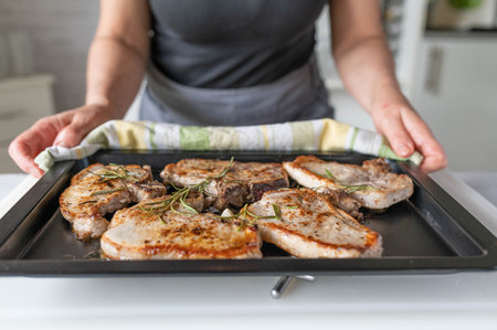 Woman hands holding baking tray with pork chops and rosemary on itの写真素材