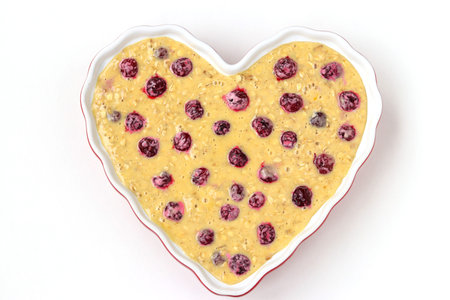 Dough for oatmeal cake with cherries in ceramic form in the shape of a heart on a white background,...
